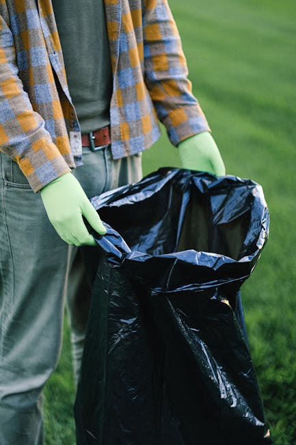 A person wearing a plaid shirt in shades of yellow, gray, and blue, along with a green long-sleeved top underneath, is seen holding open a large black rubbish bag lined with a shiny black plastic interior. The individual is standing outdoors on a grassy area, with the background showing a well-maintained lawn and blurred greenery. They are wearing light-colored trousers and a brown belt, and their hands are covered with light green gloves, indicating they are engaged in waste collection or rubbish disposal. The scene suggests a private or independent waste removal activity, possibly as part of outdoor cleaning or garden clearance. The black rubbish bag is empty and ready for waste, supporting the context of rubbish clearance services, like those offered by Rubbish Clearance Brent, which provides alternative waste management solutions beyond local authority collections. The natural lighting emphasizes the outdoor environment, and the overall composition highlights the process of rubbish handling, aligning with professional waste removal efforts.
