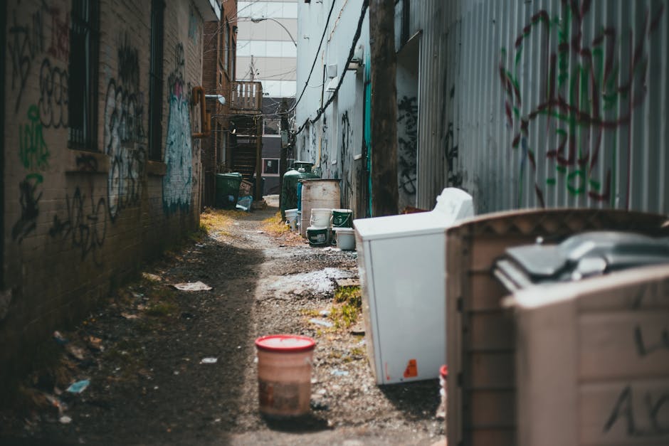 The image depicts a narrow alleyway between two urban buildings, with walls covered in colorful graffiti and peeling paint visible on the brick and metal surfaces. On the right side, a row of large, white household appliances, including a washing machine and possibly a refrigerator, are stacked haphazardly, with some partially obscured by a wooden crate. On the left, a similar arrangement of green and black waste bins and discarded items can be seen, alongside scattered rubbish and debris on the ground, which is uneven and muddy with patches of wet soil and small puddles. In the background, a small wooden staircase leading to an upper level of one building is visible, along with more graffiti, suggesting a neglected or back-of-property area often cleared through independent rubbish removal services. The lighting appears to be natural daylight, giving a subdued tone to the scene, emphasizing the cluttered, utilitarian environment typical of non-standard waste disposal sites, aligning with alternative waste handling or on-site clearance activities performed by companies like Rubbish Clearance Brent.