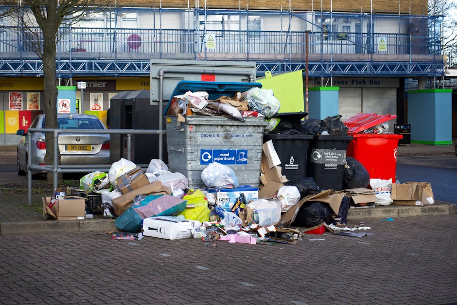 A large collection of mixed household waste and recyclables is piled on a paved sidewalk in front of a set of black, green, and red rubbish bins, with some items spilling out onto the ground. The waste includes cardboard boxes, paper bags, plastic containers, and black trash bags, some of which are ripped open, revealing their contents. Behind the debris, a grey recycling bin with symbols for mixed paper and card is visible, with its lid partially open and overflowing, containing crumpled paper, cardboard, and other refuse. To the left, a parked grey car is partially obscured by a metal railing, and in the background, there is a modern building with blue railings, large windows, and brightly colored shop fronts, suggesting an urban environment. The scene depicts an instance of informal rubbish accumulation suitable for private waste collection or rubbish removal services offered by Rubbish Clearance Brent, located near an urban station area, highlighting the need for proper waste management and clearance outside of municipal disposal systems.