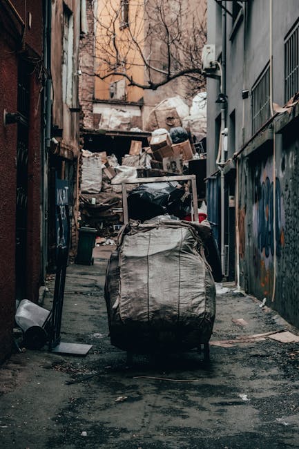 A narrow urban alleyway cluttered with various types of waste, including a large, beige duffel bag or bundle secured with straps in the foreground, positioned on the uneven, worn asphalt surface. Behind it, a collection of black and white plastic bags, some torn open, and miscellaneous discarded items are piled up against makeshift wooden pallets and cardboard boxes, creating an overflowing scene indicative of non-commercial rubbish collection. The surrounding environment consists of dimly lit brick and metal-clad walls with graffiti and peeling paint, and a few small windows and utility pipes visible on the right side. Bare trees with branches stretching across the sky can be seen in the background, above the clutter, suggesting an alley within a residential or mixed-use area, possibly used for private waste disposal or on-site clearance by waste removal services such as Rubbish Clearance Brent. The overcast lighting and general atmosphere emphasize a scene of unsorted domestic or construction waste awaiting collection or disposal, aligning with independent rubbish removal activities in urban settings.