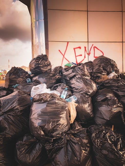 A large collection of mixed household waste and recyclables is piled on a paved sidewalk in front of a set of black, green, and red rubbish bins, with some items spilling out onto the ground. The waste includes cardboard boxes, paper bags, plastic containers, and black trash bags, some of which are ripped open, revealing their contents. Behind the debris, a grey recycling bin with symbols for mixed paper and card is visible, with its lid partially open and overflowing, containing crumpled paper, cardboard, and other refuse. To the left, a parked grey car is partially obscured by a metal railing, and in the background, there is a modern building with blue railings, large windows, and brightly colored shop fronts, suggesting an urban environment. The scene depicts an instance of informal rubbish accumulation suitable for private waste collection or rubbish removal services offered by Rubbish Clearance Brent, located near an urban station area, highlighting the need for proper waste management and clearance outside of municipal disposal systems.