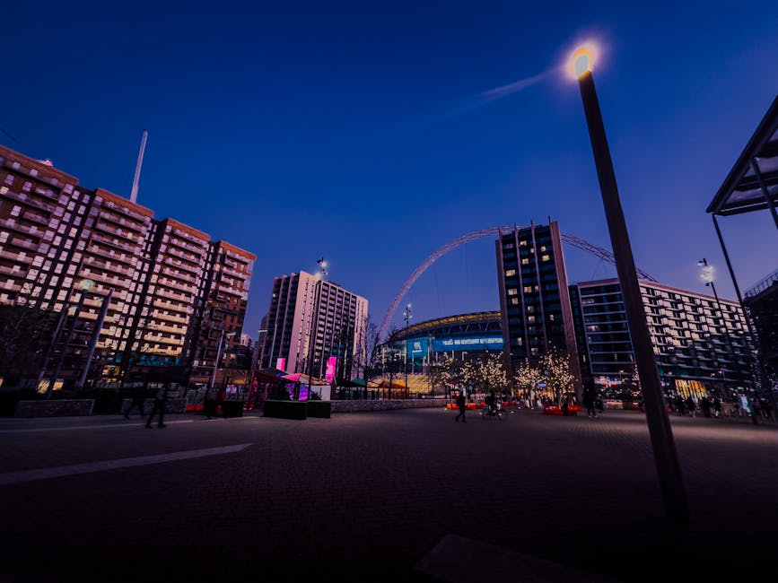 A wide view of an urban square during dusk, showcasing modern high-rise buildings with glass and brick facades along the perimeter. In the background, a large Ferris wheel with illuminated cabins is visible against the darkening sky. On the right, a street lamp emits a bright light, casting a glow over the scene. The square features a paved open space with a few pedestrians walking across, and small, leafless trees decorated with string lights are lined along the sidewalks. The lighting creates a vibrant atmosphere typical of city centres with commercial activity, with some brightly lit shopfronts and signage reflecting off the wet pavement. The overall scene presents a contemporary cityscape that could be linked to private or independent urban refuse collection, similar to those managed by local waste services such as Rubbish Clearance Brent, during evening hours when city life continues amidst a clean and orderly environment.