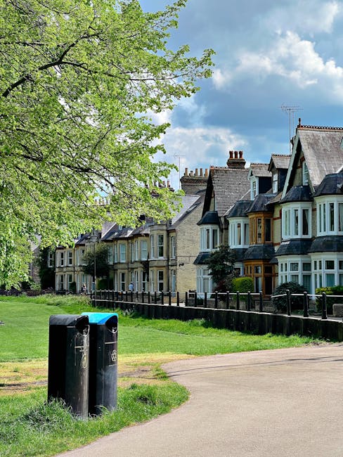 A public park area in a residential neighbourhood featuring two black bollard-style rubbish bins positioned on a grassy verge near a paved pathway. In the background, there is a row of terraced Victorian-style houses with bay windows, brick and stucco facades, some with decorative woodwork and chimneys visible against a blue sky with scattered clouds. A large tree with light green leaves extends its branches over the scene, providing partial shade. The lawn surrounding the bins is well-maintained, and a low black metal fence runs parallel to the houses, separating the green space from the residential properties. The image captures the peaceful atmosphere typical of a suburban environment that may benefit from private waste collection services as an alternative to local authority rubbish removal, with visible emphasis on the urban landscape, planting, and waste disposal points for on-site clearance or private rubbish management. The overall lighting suggests a bright, clear day with natural daylight illuminating the scene.